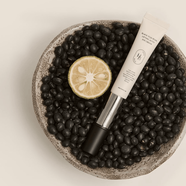 Black Yuja Bean Lifting Auto Eye Cream liggande i en skål fylld med svarta bönor och en halverad citrusfrukt – en visuell representation av produktens naturliga ingredienser.