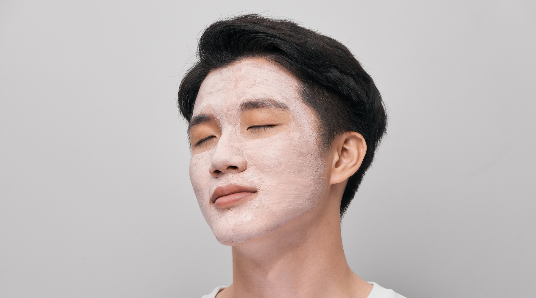 Asiatisk man med avslappnat ansikte och vit ansiktsmask mot en ljusgrå bakgrund.