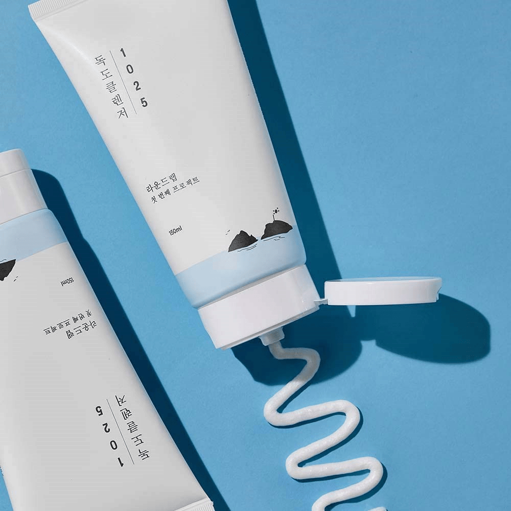To tuber af 1025 Dokdo Cleanser på blå baggrund, hvoraf en med åben prop og en bølget streng rengøring presset ud.