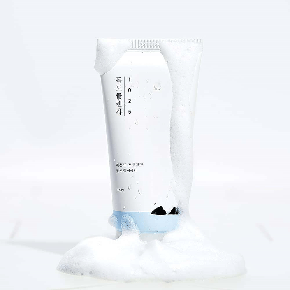 1025 Dokdo Cleanser i hvid tube dækket af skum, med vandperler og skum rundt om bunden, frilagt mod lys baggrund.