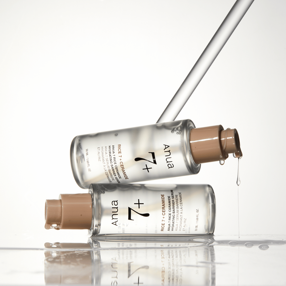 Billedet viser to gennemsigtige flasker af "Anua 77 Rice + Ceramide Hydrating Barrier Serum" med dråber, der løber fra en pipette. Serummet, som er beriget med ris og ceramider, er beregnet til at fugte og styrke hudens barriere. Flaskerne er elegant designet, hvilket forstærker produktets premiumfølelse.