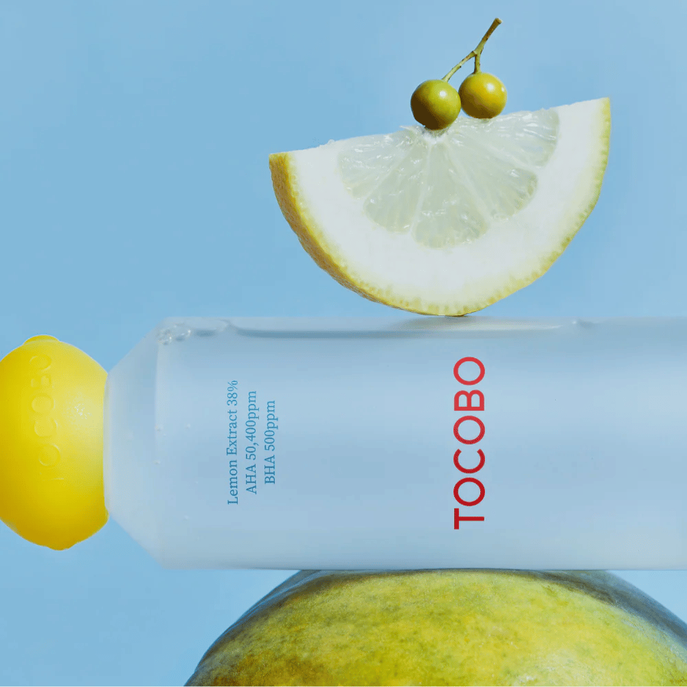 Flaske med TOCOBO AHA BHA Lemon Toner, dekoreret med et låg i form af en citron. En citronskive og små grønne bær balancerer ovenpå flasken mod en blå baggrund.