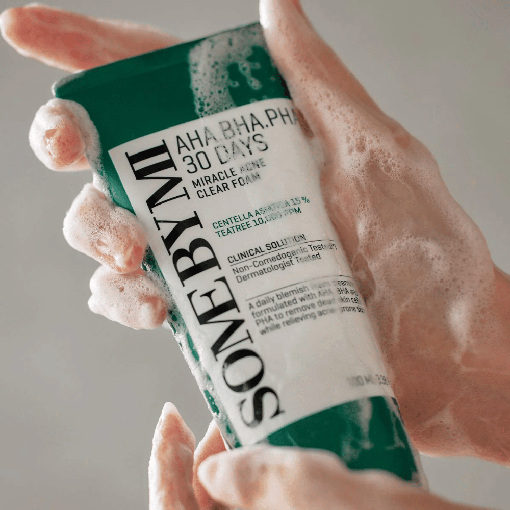 En person holder en tube af "SOME BY MI AHA, BHA, PHA 30 Days Miracle Acne Clear Foam" med skummende indhold over hænderne. Produktet er beregnet til daglig brug for at bekæmpe akne og rense porerne med dets aktive ingredienser som centella, tea tree olie og syrer som AHA, BHA og PHA.