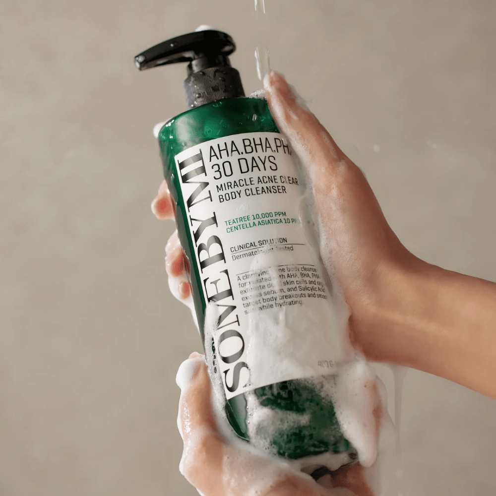 En person holder en flaske SOME BY MI AHA.BHA.PHA 30 Days Miracle Acne Clear Body Cleanser. Flasken er grøn med en hvid etiket og er dækket af skum fra rengøringsmidlet. Personen vasker sig med produktet, mens vand løber over hænderne.