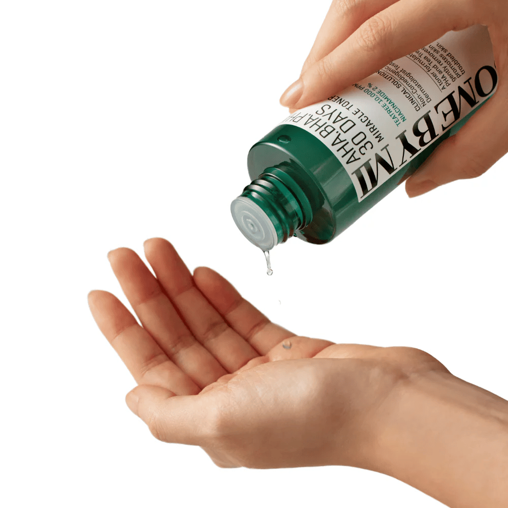 En hånd holder en grøn flaske med teksten "AHA, BHA, PHA 30 Days Miracle Toner" og presser en klar væske ud i den anden hånds håndflade, klar til brug.