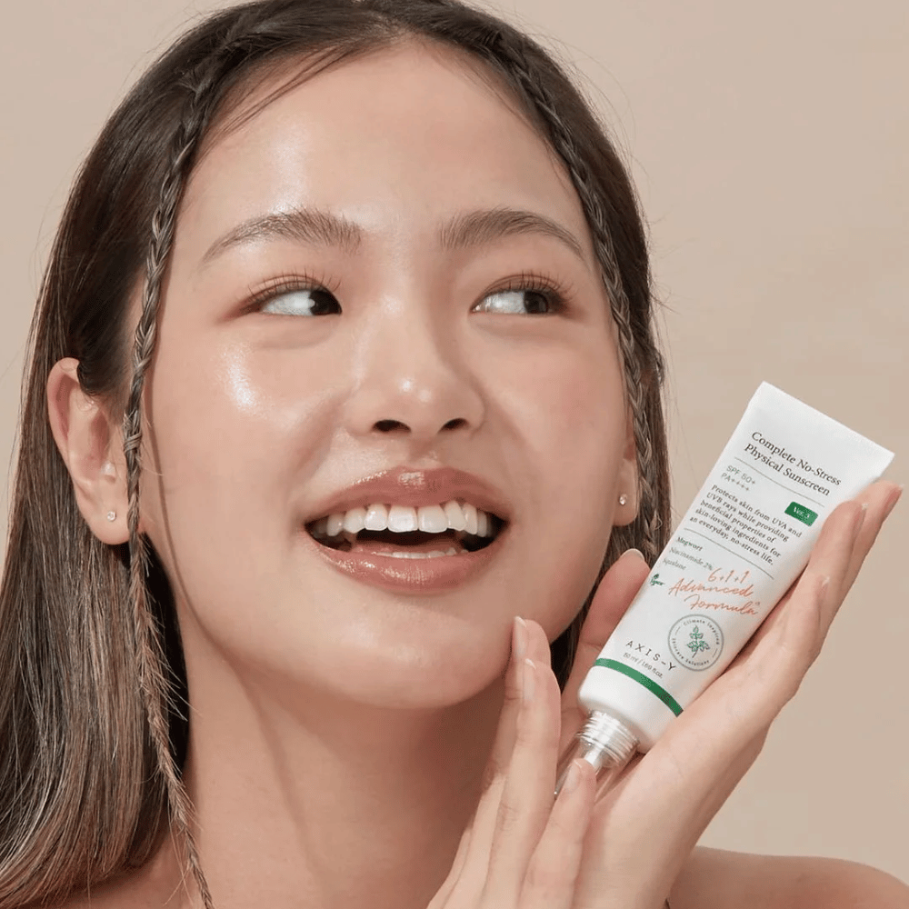 Billedet viser en ung kvinde, der smiler bredt og holder en tube med AXIS-Y Complete No-Stress Physical Sunscreen. Hun har et strålende og naturligt udseende med minimal makeup, hvilket fremhæver produktet, hun præsenterer.