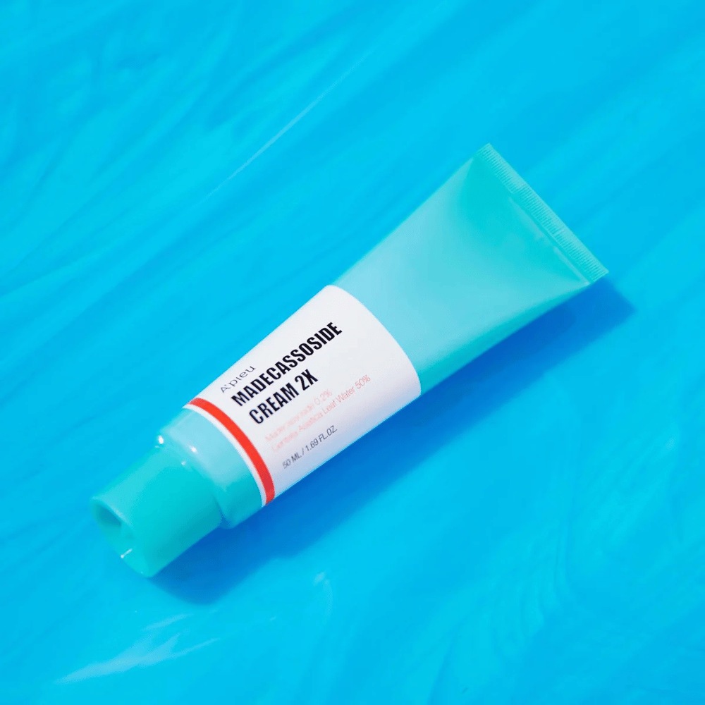 En tube A'PIEU Madecassoside Cream 2X ligger diagonalt over en blå baggrund med vandlignende rynker. Tuben har en hvid krop med en lys turkis hætte og en rød til orange farvet etiket, hvor produktnavnet og den aktive ingrediens Madecassoside 2% er tydeligt skrevet. På etiketten står også mængden, som er 50 ml / 1.8 fl oz. Den lyse og farverige præsentation giver en frisk og energisk følelse.