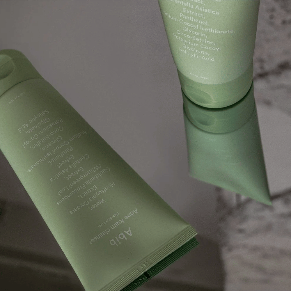 Grøn tube af Abib Acne Foam Cleanser Heartleaf liggende på et spejlbord med produktets forside og tekst spejlet. Ingredienser som salicylsyre, centella og hjertbladsekstrakt ses tydeligt på emballagen.