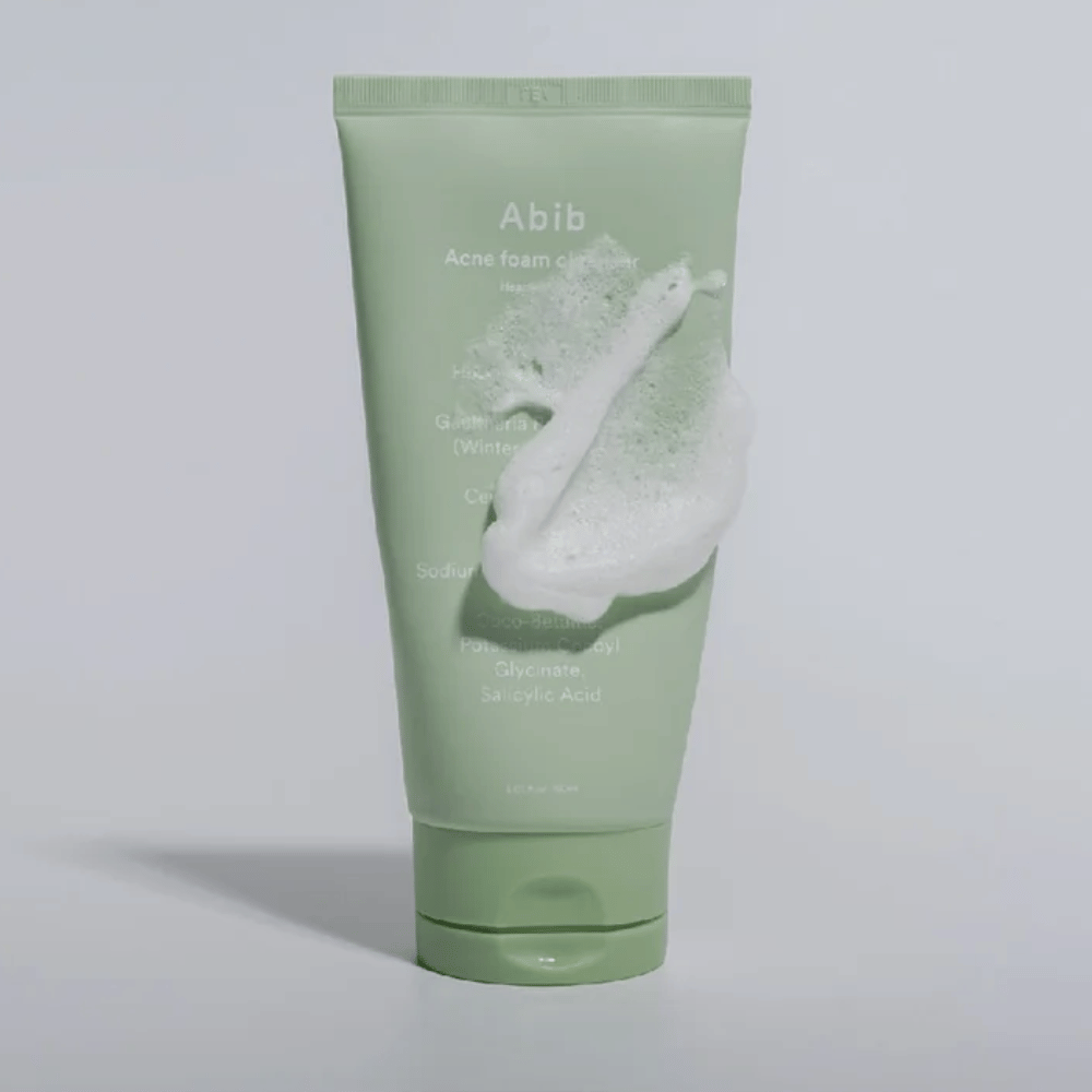 Grøn tube med Abib Acne Foam Cleanser Heartleaf, dækket af et hvidt skum som illustrerer den skummende renseeffekt. Produktet indeholder hjertbladsekstrakt og salicylsyre til akne-udsat hud.