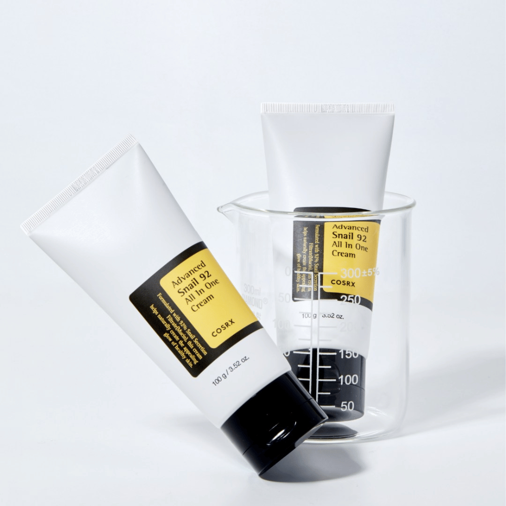 Et rør af COSRX Advanced Snail 92 All in One Cream liggende ved siden af en omvendt gennemsigtig målecylinder på en hvid baggrund.