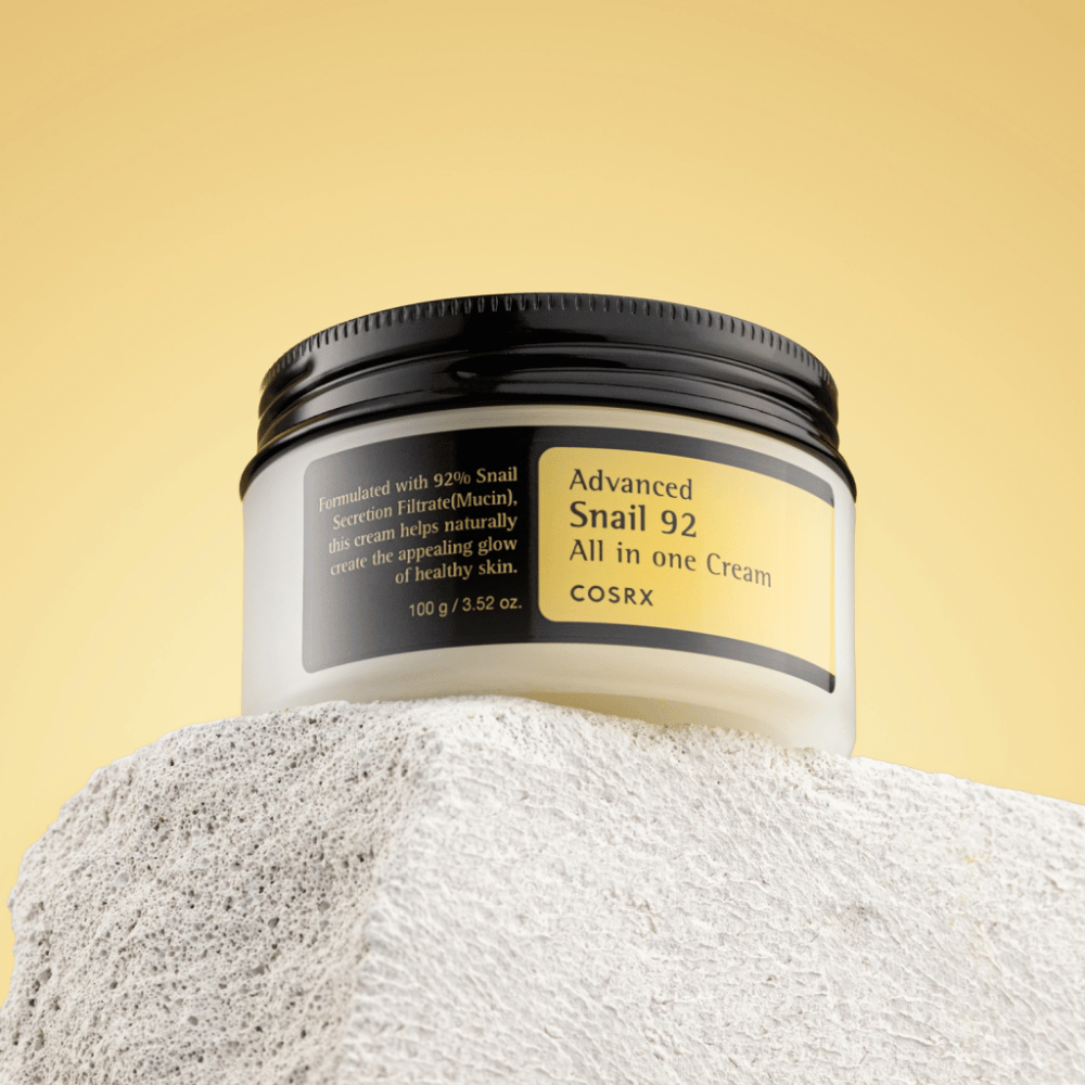 En krukke af COSRX Advanced Snail 92 All in one Cream placeret på en stenoverflade med en gul baggrund.