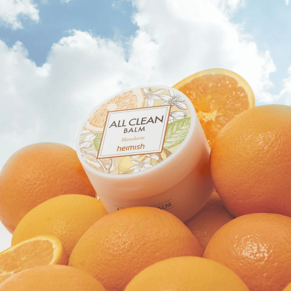Dåse af Heimish All Clean Balm Mandarin placeret på en bunke appelsiner, med en blå himmel i baggrunden.