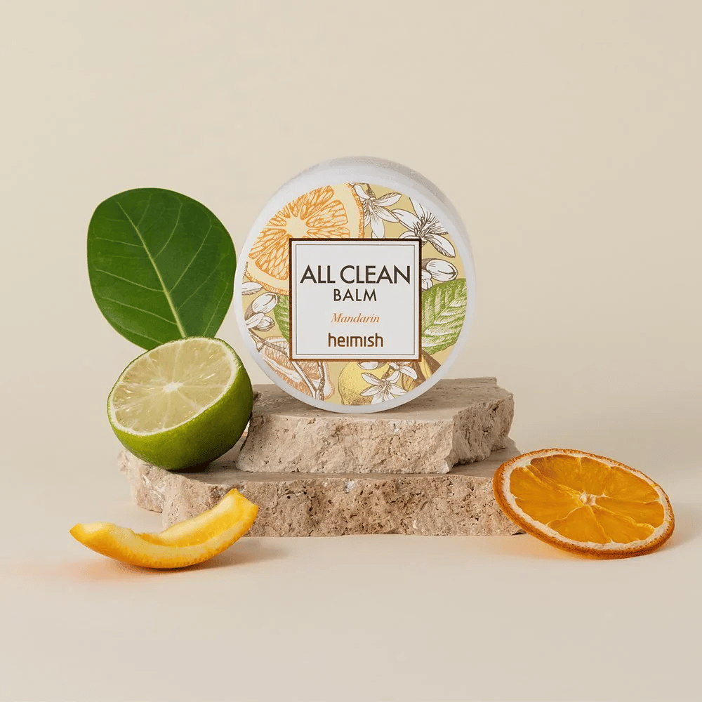 Dåse af Heimish All Clean Balm Mandarin placeret på sten, omgivet af mandarinskræl og citrusblade, foran en lys baggrund.
