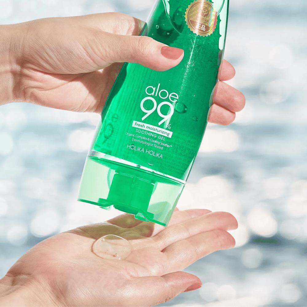 En hånd holder en grøn tube med Holika Holika Aloe 99% Soothing Gel, mens en anden hånd fanger en klat gel, der dispenseres. Baggrunden er sløret og viser et vandigt, glitrende mønster.