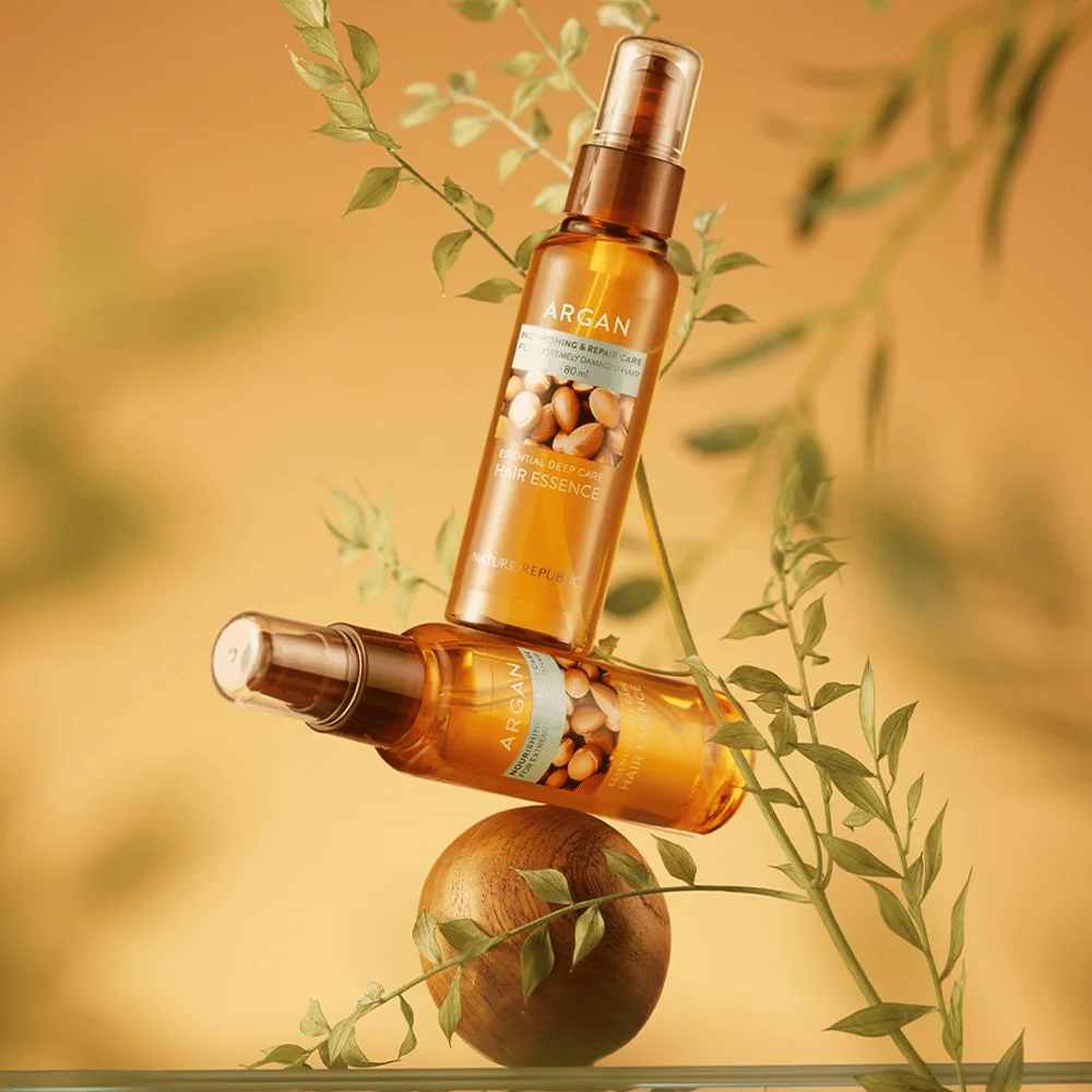 To flasker Nature Republic Argan Essential Moisture Hair Oil placeret blandt grønne blade – silkeagtig glans til tørt hår.