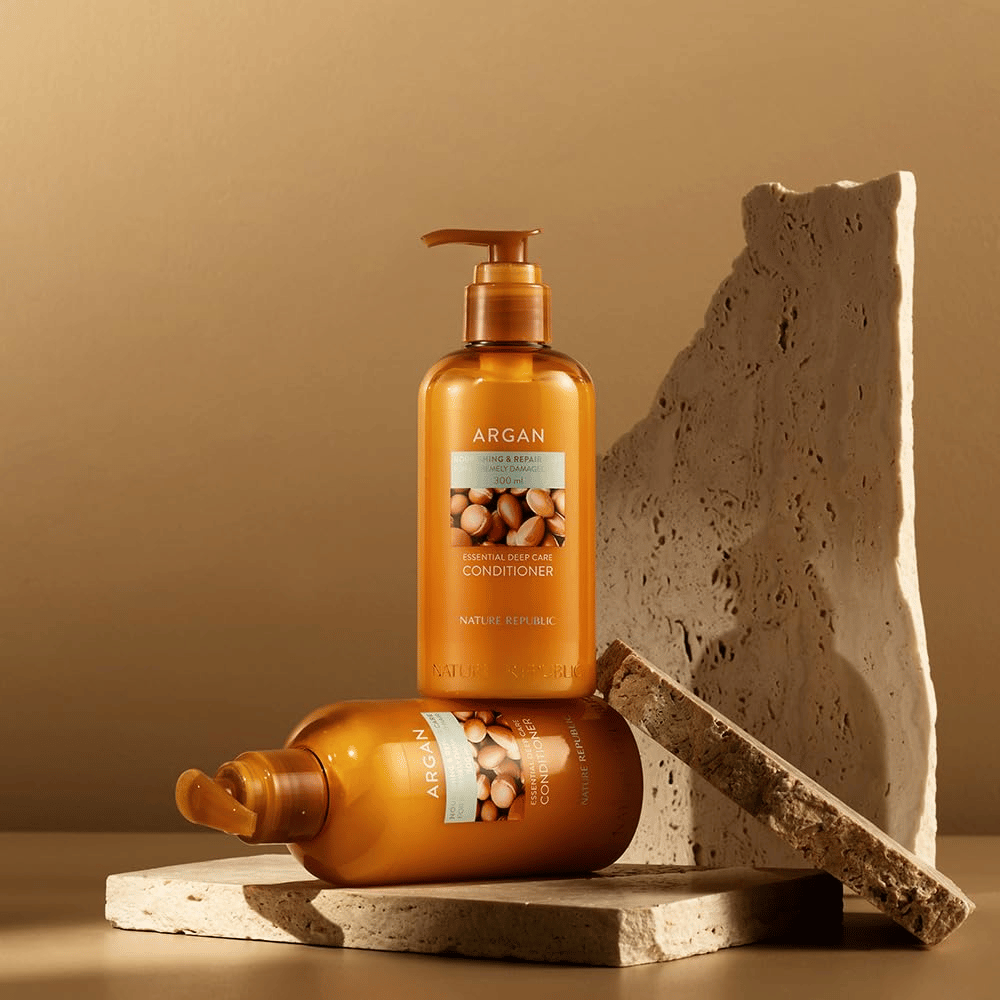 To flasker af Nature Republic Argan Essential Moisture Repairing Conditioner på stenunderlag i varm tone.