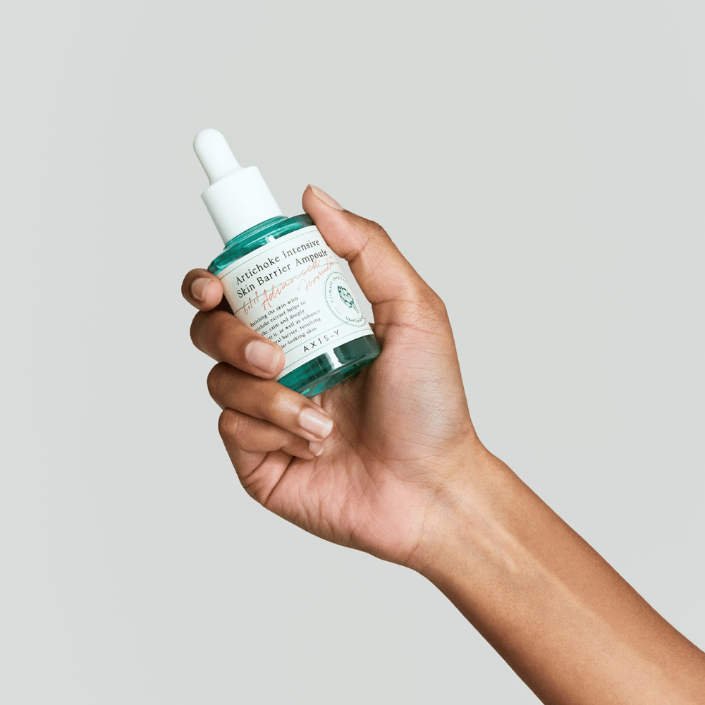 En hånd holder en flaske hudplejeserum med etiketten "Artichoke Intensive Skin Barrier Ampoule.