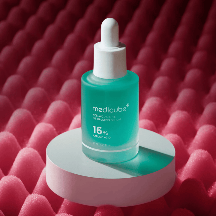 Produktbillede af Medicube Azelaic Acid 16 BB Calming Serum placeret på hvid podieoverflade med rosa tekstureret baggrund, som fremhæver serumets beroligende og hudforbedrende profil.