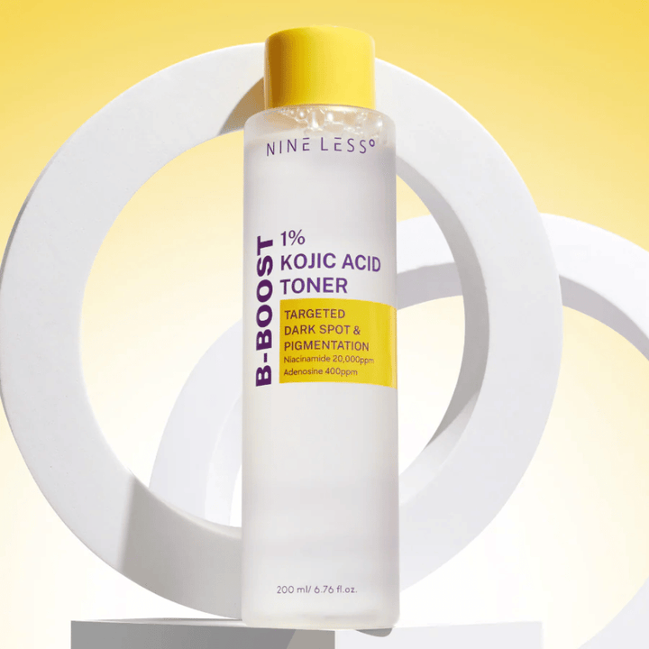 NINE LESS B-Boost 1% Kojic Acid Toner stående mot gul bakgrund med texten "Targeted Dark Spot & Pigmentation". Hudton-utjämnande toner med niacinamid och adenosin.