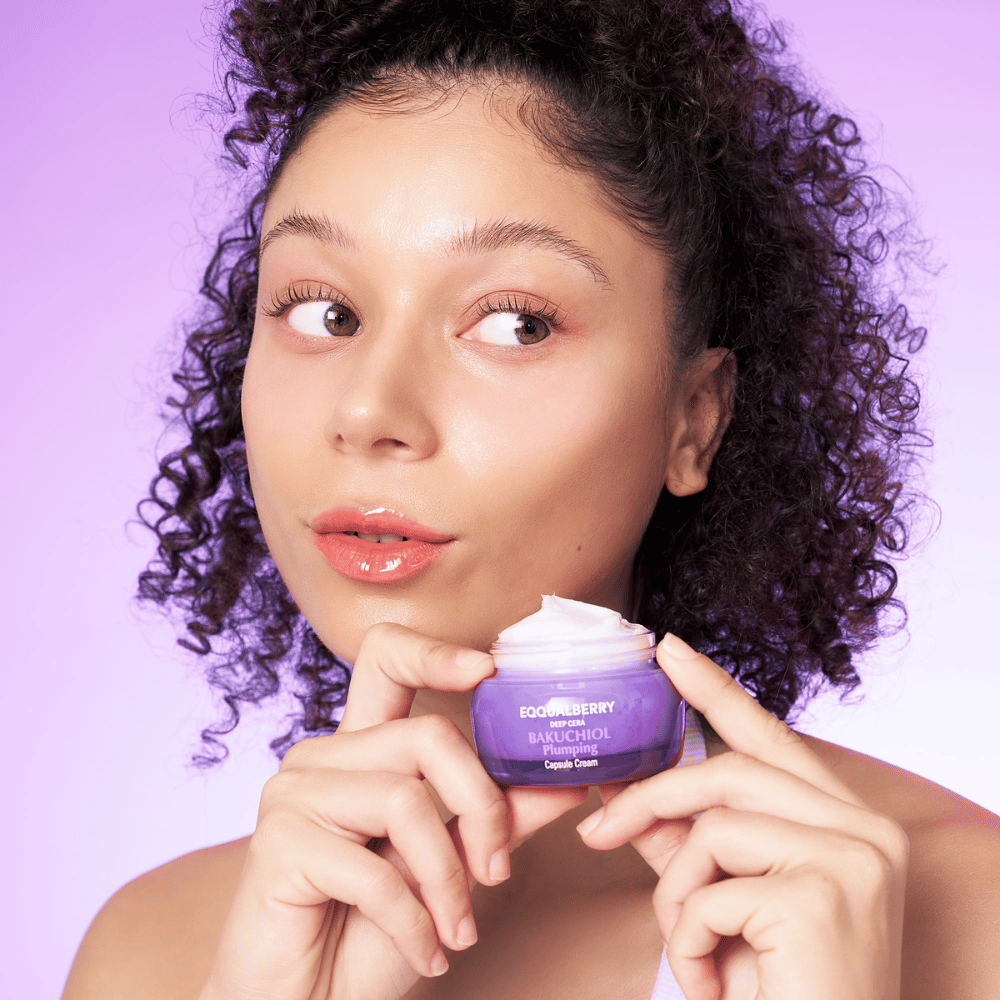 Kvinde holder EQQUALBERRY Bakuchiol Plumping Capsule Cream i hånden og viser sin fugtede, strålende hud mod en lilla baggrund.