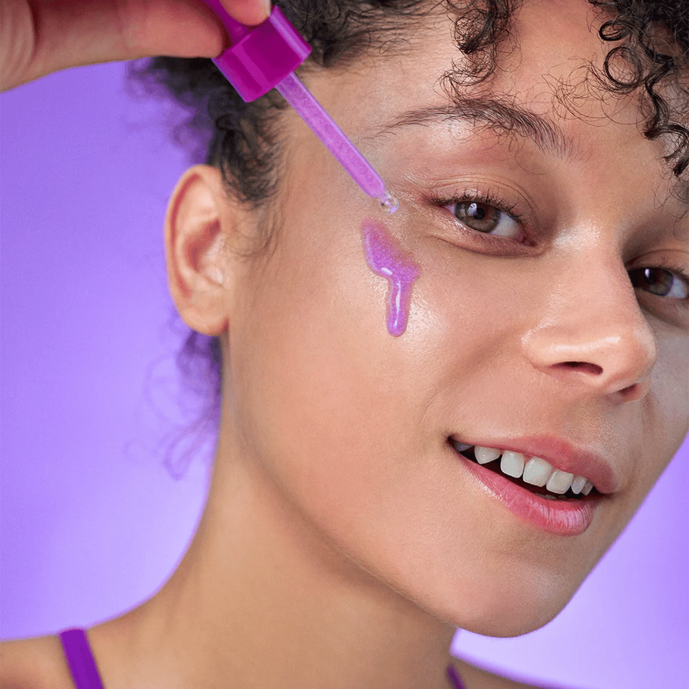 Smilende kvinde påfører et lilla skinnende serum på huden med en pipette. Strålende hud og lilla baggrund, der matcher produktets farvetone.