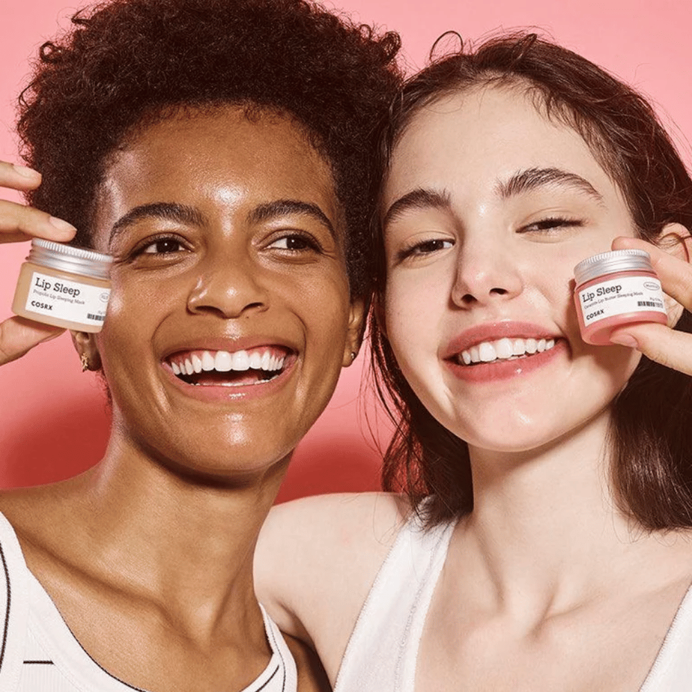 To personer smiler og holder op COSRX Lip Sleep-krukker. Billedet formidler bløde, fugtede læber og produktets naturlige glødgivende effekt.