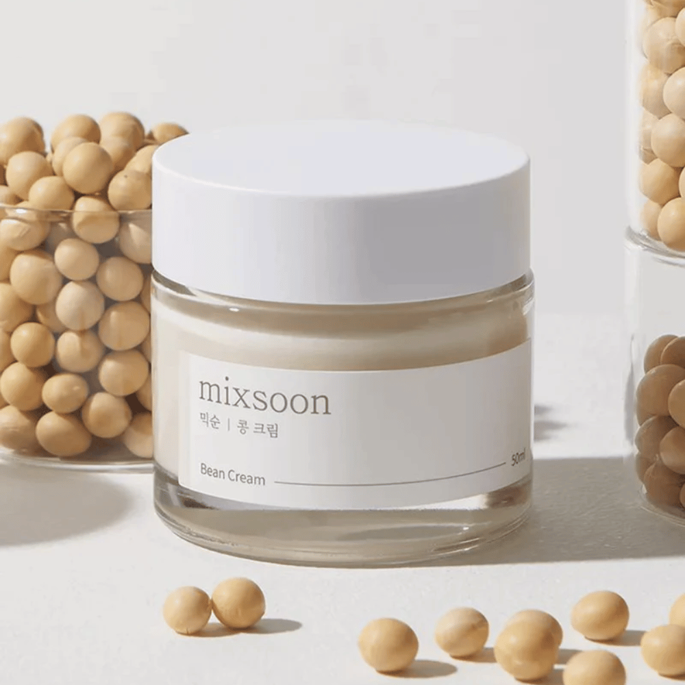 En gennemsigtig krukke med hvid etiket og låg, mærket "mixsoon 팥 크림" og "Bean Cream" med en angivet volumen på "50ml". Krukken indeholder en lysegul creme og er placeret på en lys overflade. Ved siden af krukken står en glasbeholder fuld af lysebrune bønner, der er spildt ud på overfladen, hvilket antyder, at cremen indeholder bønneekstrakt. Baggrunden og belysningen giver en varm og naturlig atmosfære.