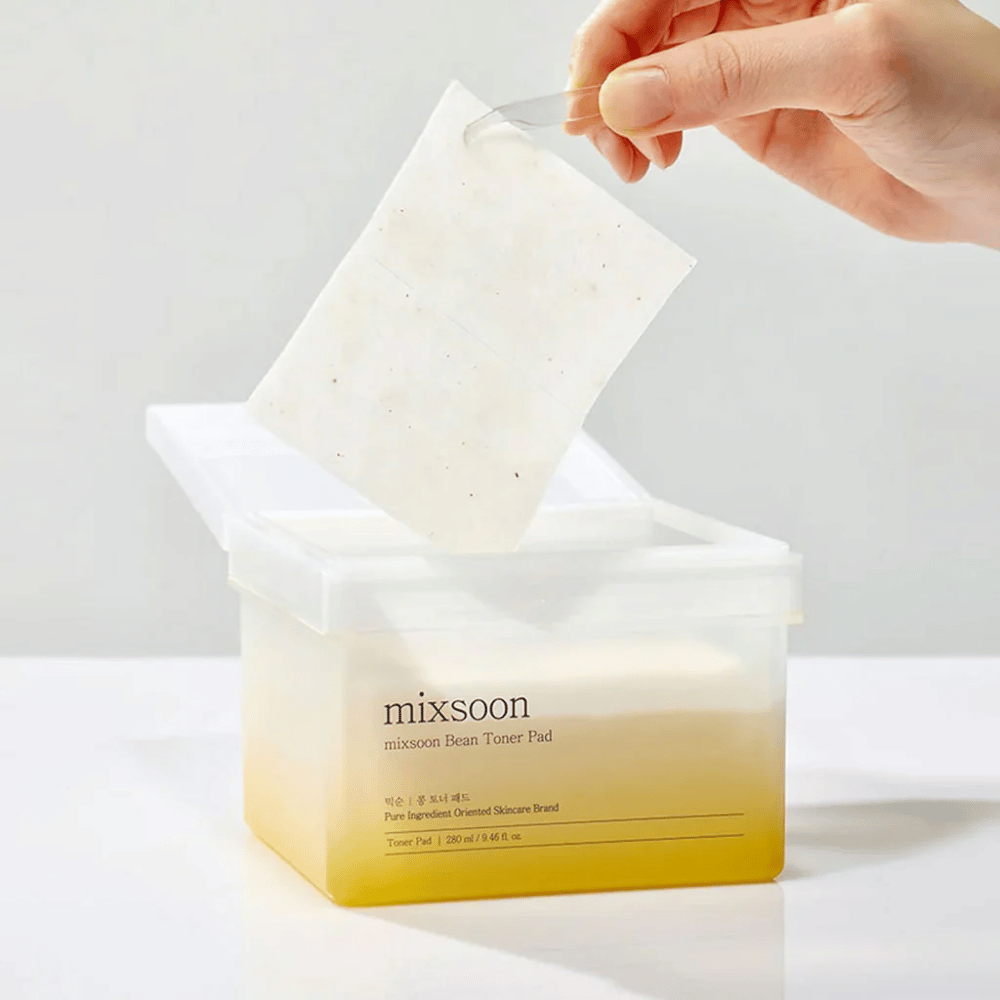 En hånd holder en pincet og løfter en Mixsoon Bean Toner Pad fra en firkantet, gennemsigtig beholder med en gullig tone. Beholderen indeholder 70 toner pads og har tekst, der beskriver produktet.