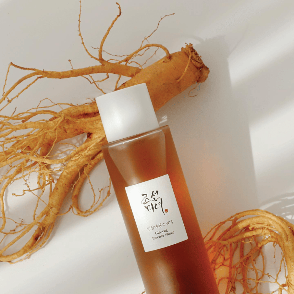 En brun flaske Beauty of Joseon Ginseng Essence Water med hvid kork ligger ved siden af en frisk ginsengrod på hvid baggrund.
