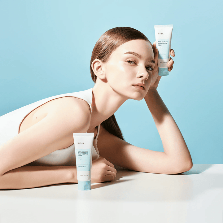 Livsstilsbillede, hvor en model poserer med iUNIK Beta-Glucan Daily Moisture Cream mod en lyseblå baggrund. Billedet formidler en ren, frisk og fugtgivende følelse, samtidig med at produktet eksponeres tydeligt.