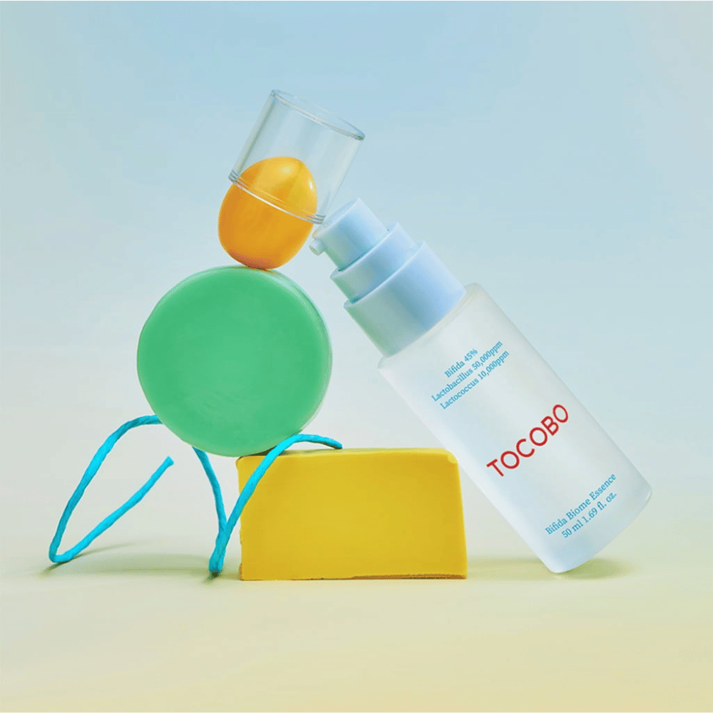 TOCOBO Bifida Biome Essence i mat pumpflaske med rød logo, placeret mod farverig baggrund med grønne, gule og orange geometriske former. Tekst på flasken angiver Bifida 5 %, Lactobacillus 3 000 ppm og Lactococcus 1 000 ppm.