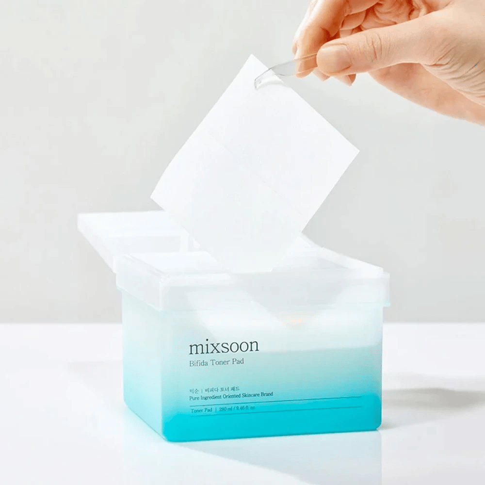 En transparent beholder med en blå og hvid gradient, der indeholder Bifida toner pads. En hånd tager en toner pad fra beholderen, som er mærket med "mixsoon Bifida Toner Pad" og rummer 280 ml (9,46 fl. oz.) toner pads beregnet til hudpleje.