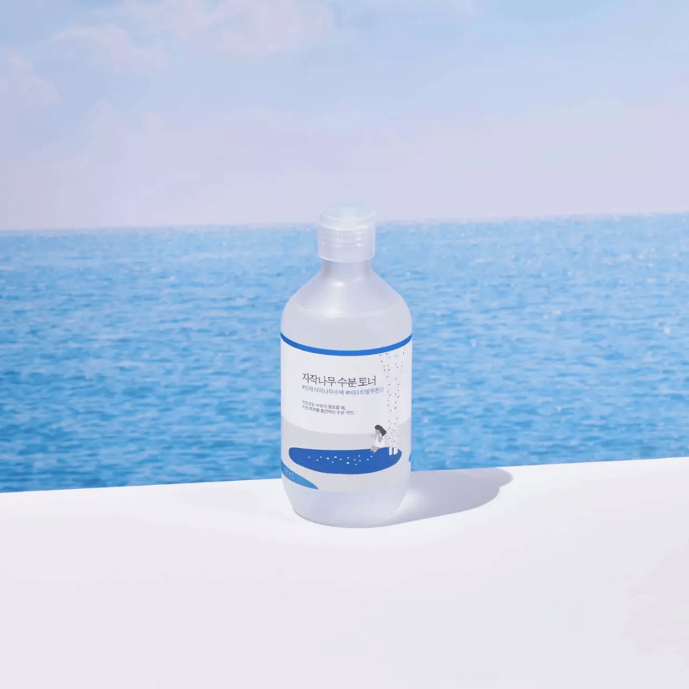 Flaske af ROUND LAB Birch Juice Moisturizing Toner står på en hvid overflade med hav og blå himmel i baggrunden
