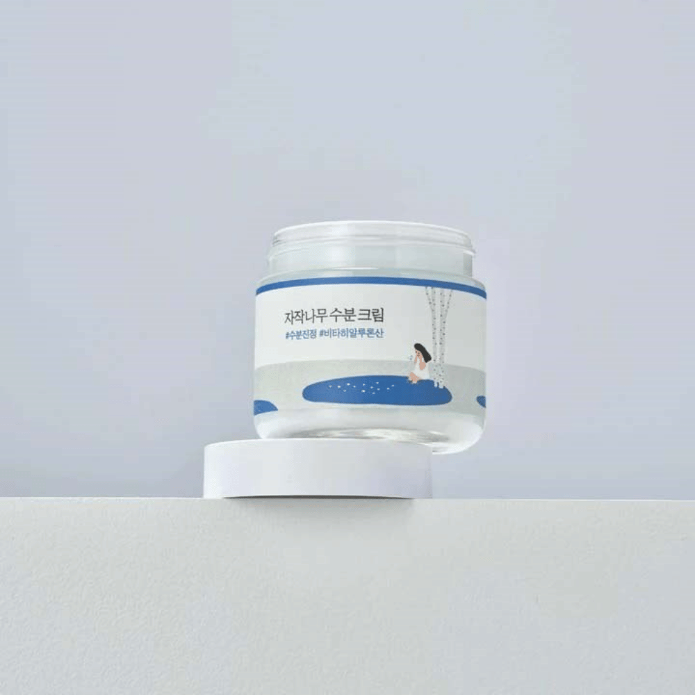 Round Lab Birch Juice Moisturizing Cream i gennemsigtig krukke med hvid etiket og blå illustration af kvinde ved birk, stående på hvid overflade mod lys baggrund.