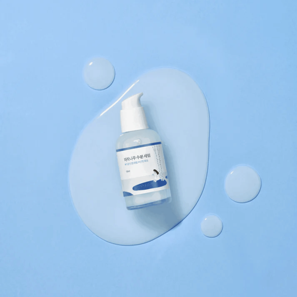 Round Lab Birch Juice Moisturizing Serum i gennemsigtig pumpeflaske placeret på en blå baggrund med dråber og spildt serum.