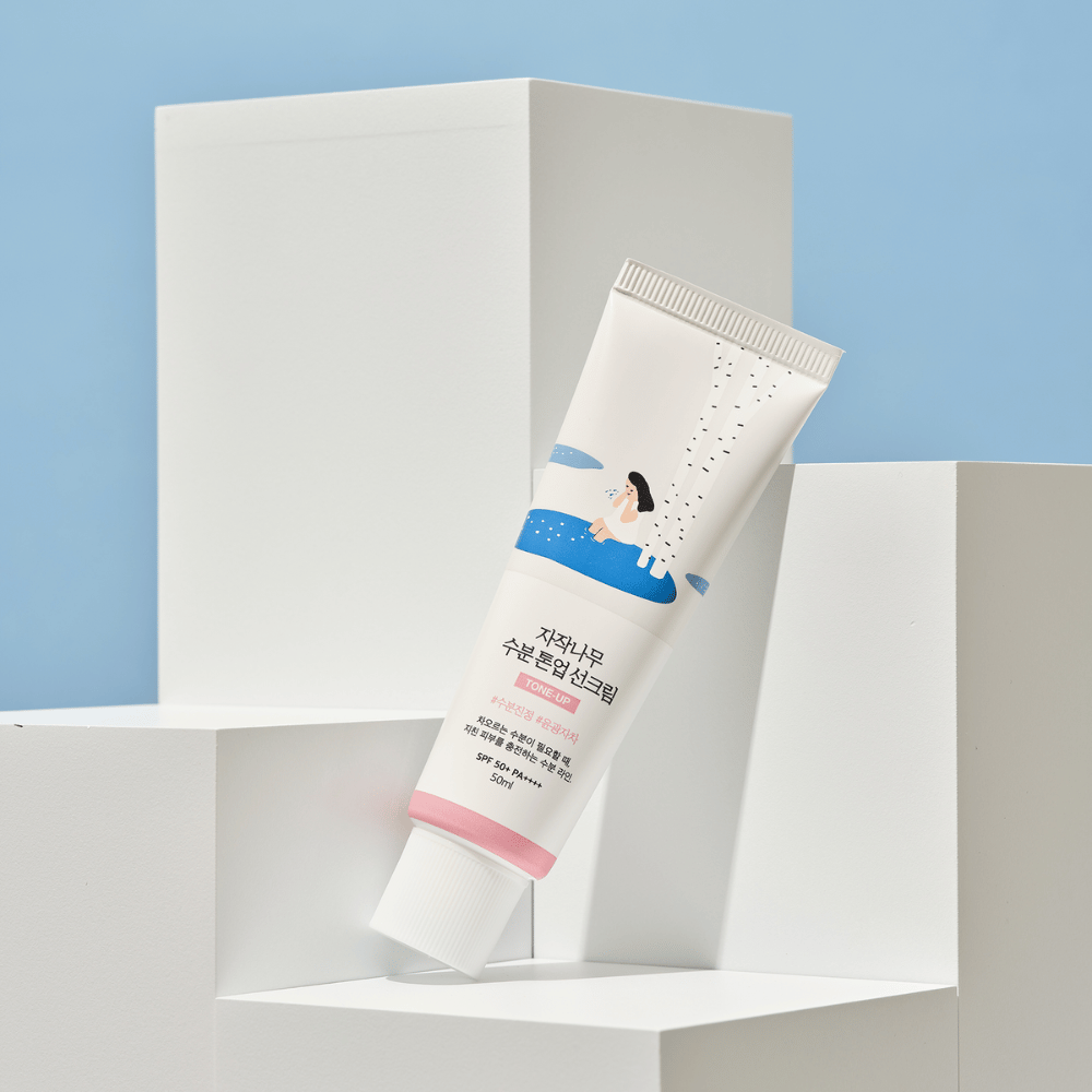 En tube med Birch Juice Moisturizing Tone Up Sunscreen placeret på hvide kuber mod en lyseblå baggrund. Tuben er hvid med en illustration af en kvinde ved en birk og vand. Teksten på koreansk angiver, at produktet har SPF 50+ PA++++ og indeholder 50 ml.