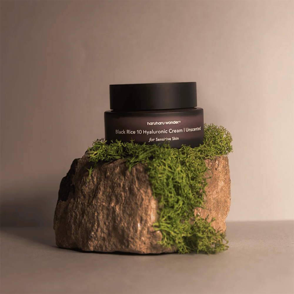 Billedet viser en bøtte hudcreme mærket "haruharu wonder Black Rice 10 Hyaluronic Cream Unscented for Sensitive Skin". Bøtten er sort og står på en stor sten dækket med lysegrøn mos, hvilket skaber en naturlig og jordnær følelse.