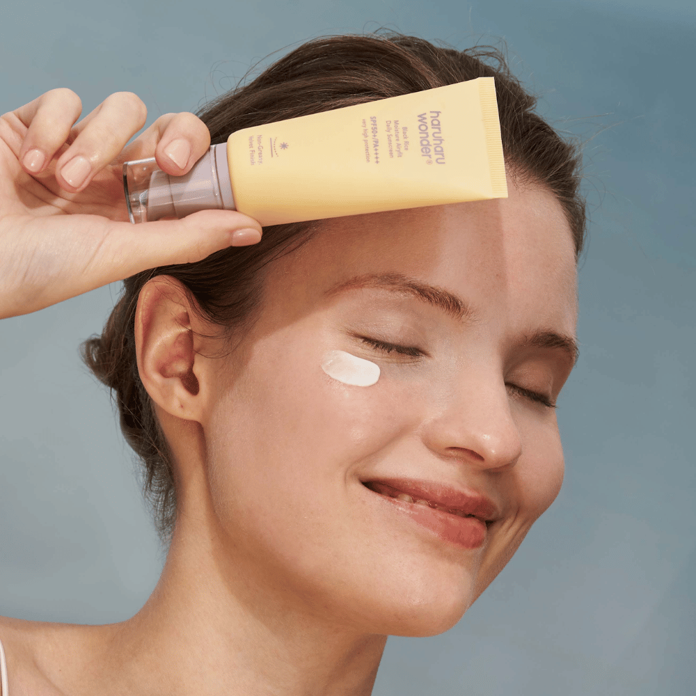 En smilende kvinde påfører Black Rice Moisture Airyfit Daily Sunscreen SPF 50+ PA++++ fra Haruharu Wonder på sit ansigt. Hun holder en gul tube med solcreme og har en klat af produktet på kinden. Baggrunden er lyseblå.