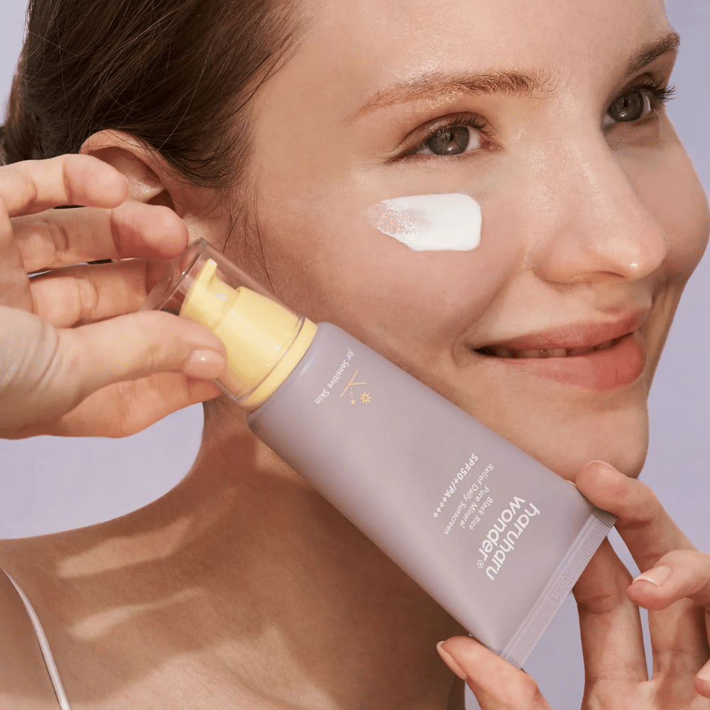 En smilende kvinde påfører Haruharu Wonder Black Rice Pure Mineral Relief Daily Sunscreen SPF 50+ PA++++ på sit ansigt. Hun holder en lilla tube med solcreme og har en klat af produktet på kinden. Baggrunden er lys lilla.