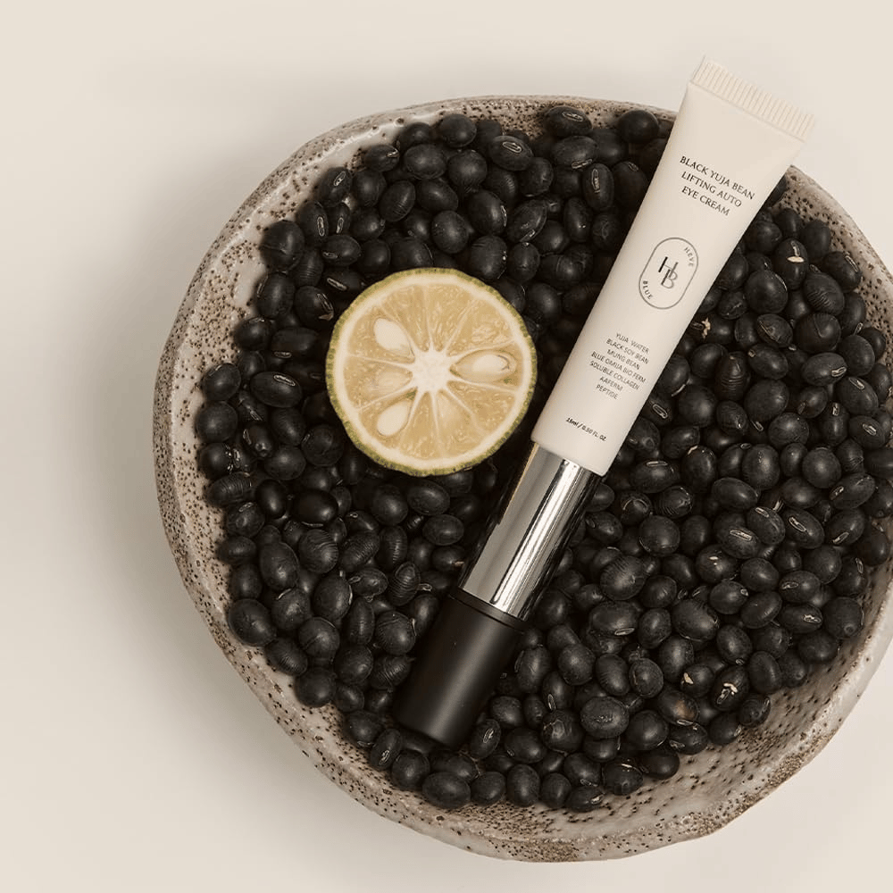 Black Yuja Bean Lifting Auto Eye Cream liggende i en skål fyldt med sorte bønner og en halveret citrusfrugt – en visuel repræsentation af produktets naturlige ingredienser.
