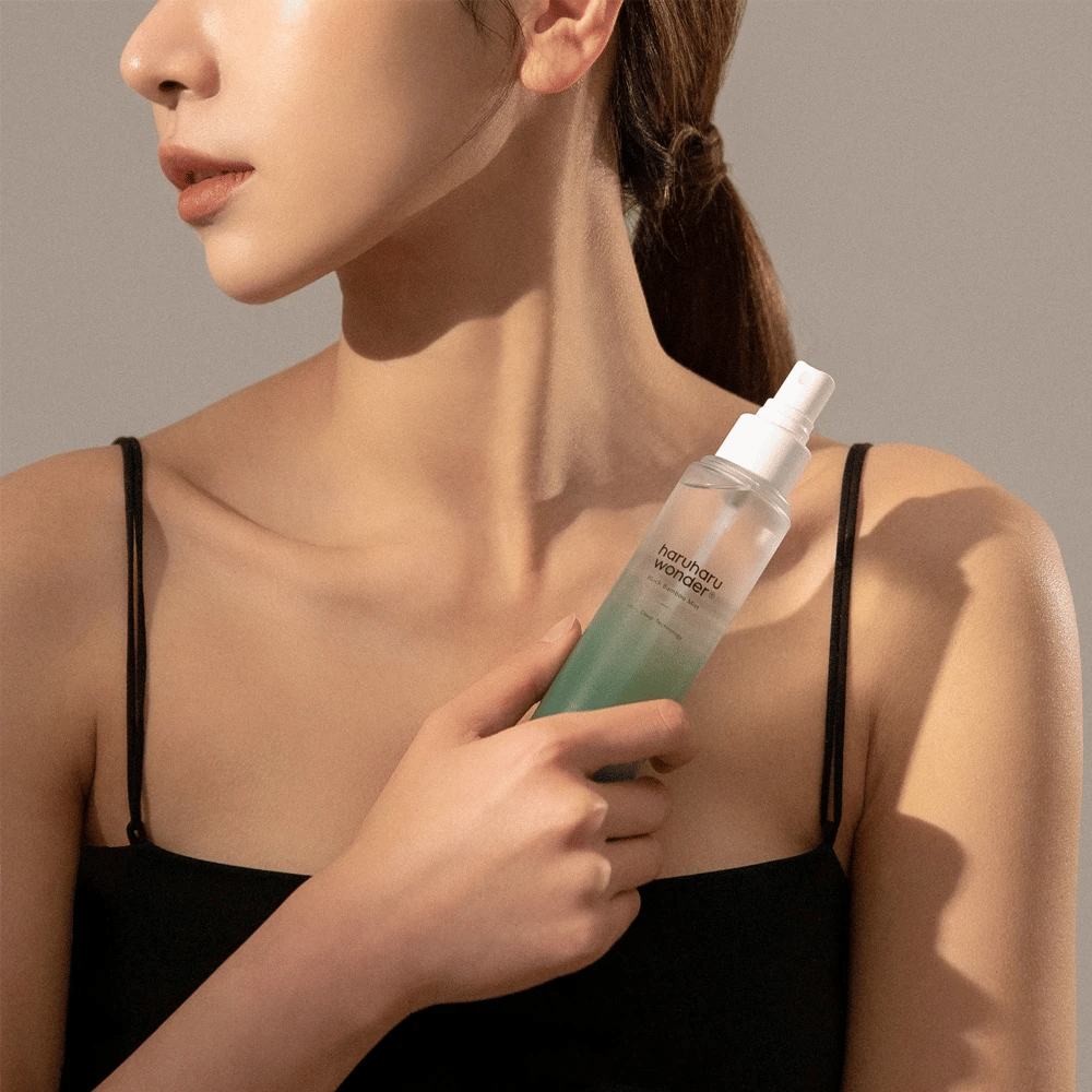 Billede af HaruHaru Wonder Black Bamboo Mist der holdes af en kvinde. Produktet er i en transparent sprayflaske med en grøn tone og en hvid pumpe. Kvinden har et afslappet look, bærer en sort top og fokuserer på at bruge misten til at give huden fugt og glød. Flasken er en del af en hudplejerutine.