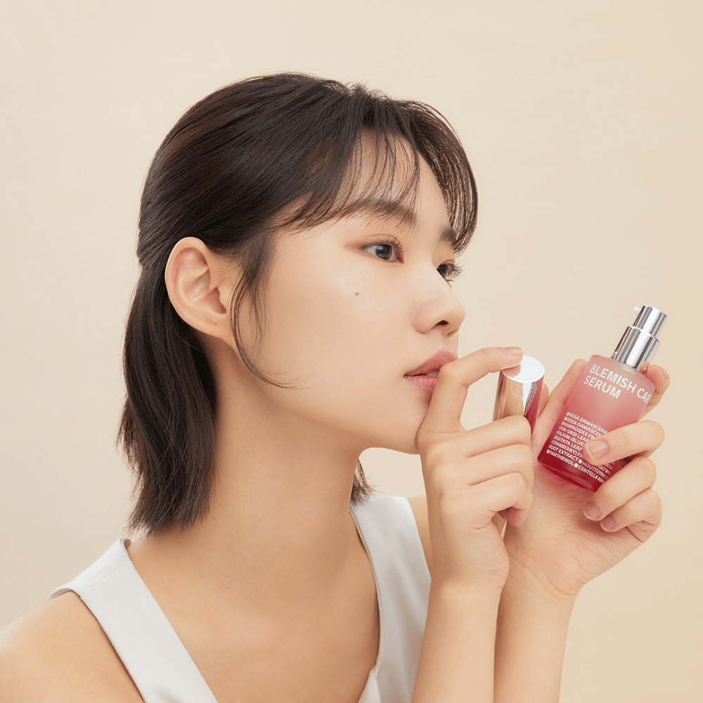 En kvinde med mørkt hår holder en flaske Blemish Care Serum i den ene hånd og tager låget af med den anden. Hun har et neutralt ansigtsudtryk, og baggrunden er lys beige, hvilket giver et blødt og minimalistisk indtryk.