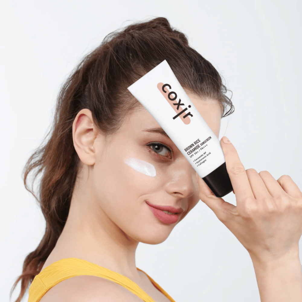 Kvinde med solcreme på kinden holder en tube Coxir Brown Rice Ceramide Sunscreen, SPF 50+ PA+++, mod panden og smiler let.