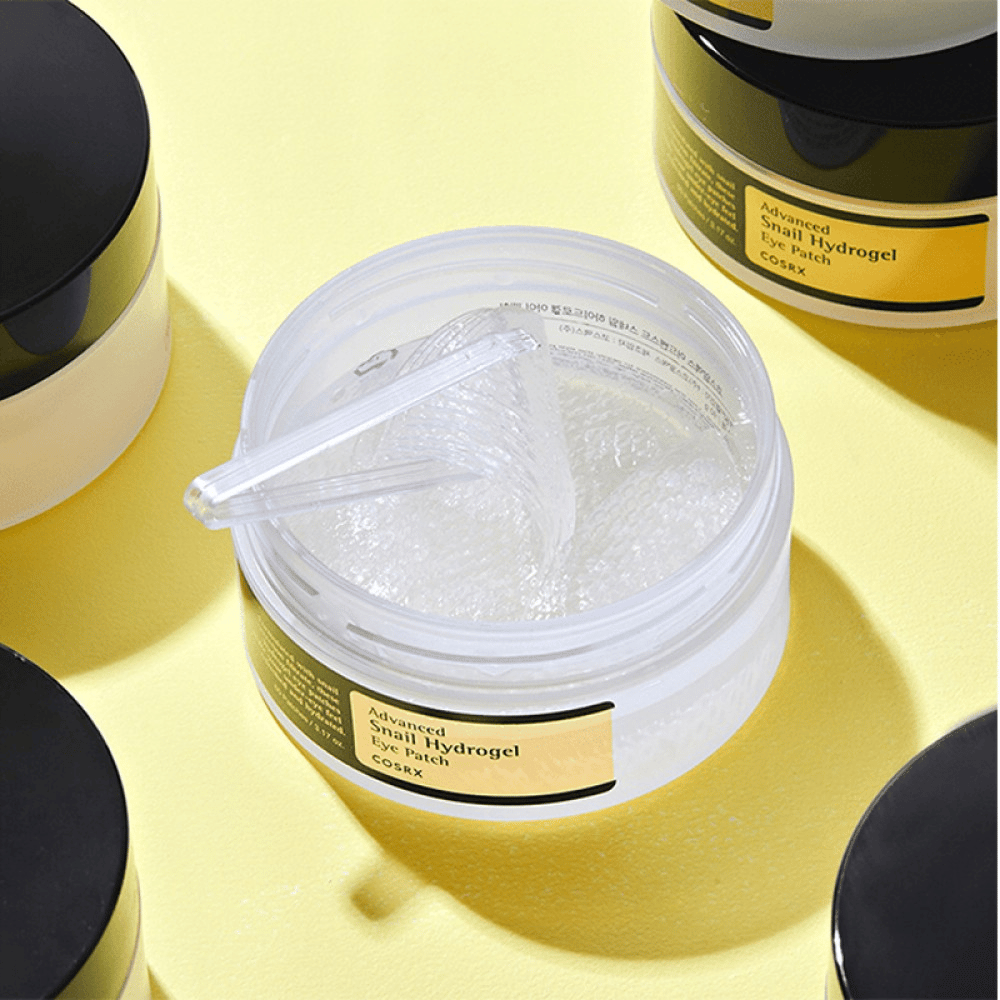 En åben krukke COSRX Advanced Snail Hydrogel Eye Patch med gennemsigtige øjenplastre og en lille spatel, omgivet af flere lukkede krukker på gul baggrund.
