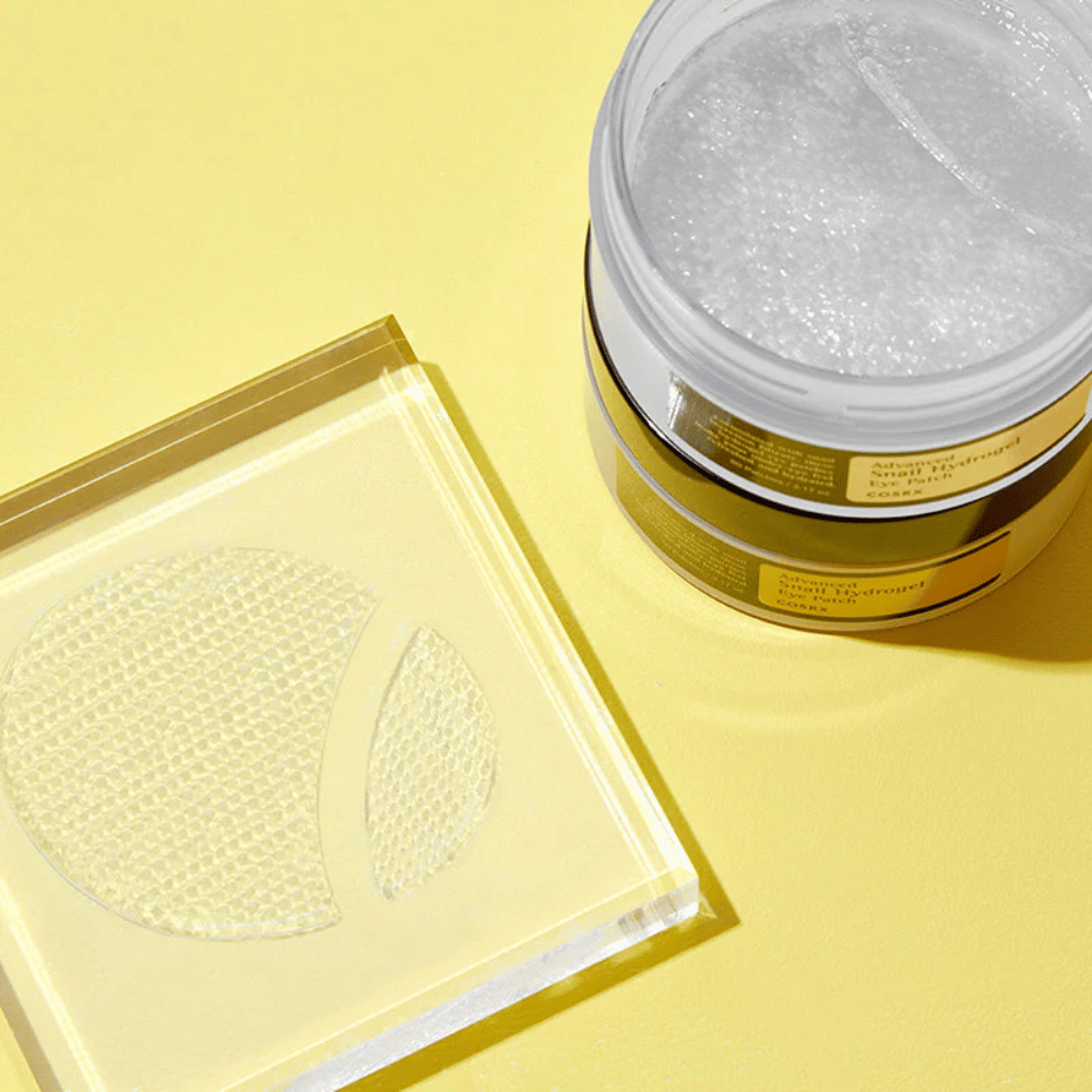 En åben krukke COSRX Advanced Snail Hydrogel Eye Patch står på gul baggrund ved siden af en gennemsigtig bakke, hvor to øjenplastre er placeret.