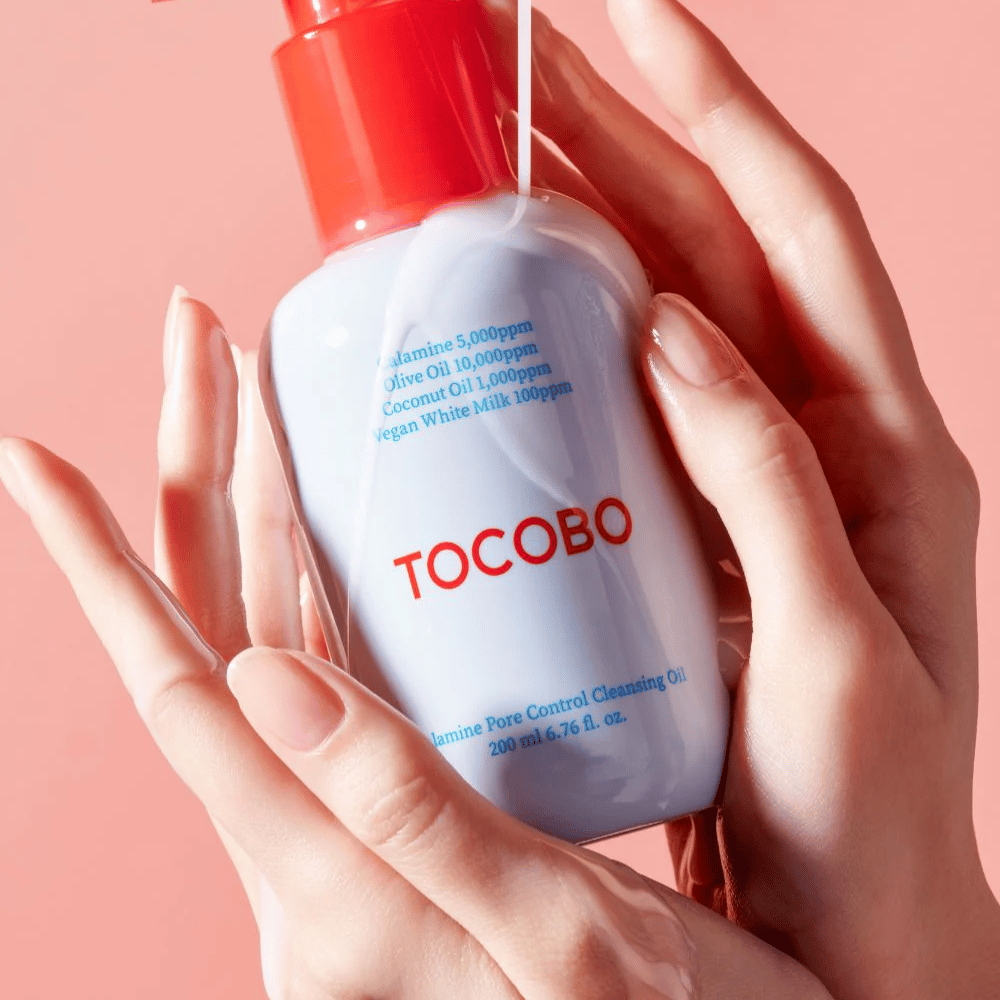 TOCOBO Calamine Pore Control Cleansing Oil holdes i hænder mod en lyserød baggrund, flaskens røde pumpe og ingrediensliste er tydeligt synlige.