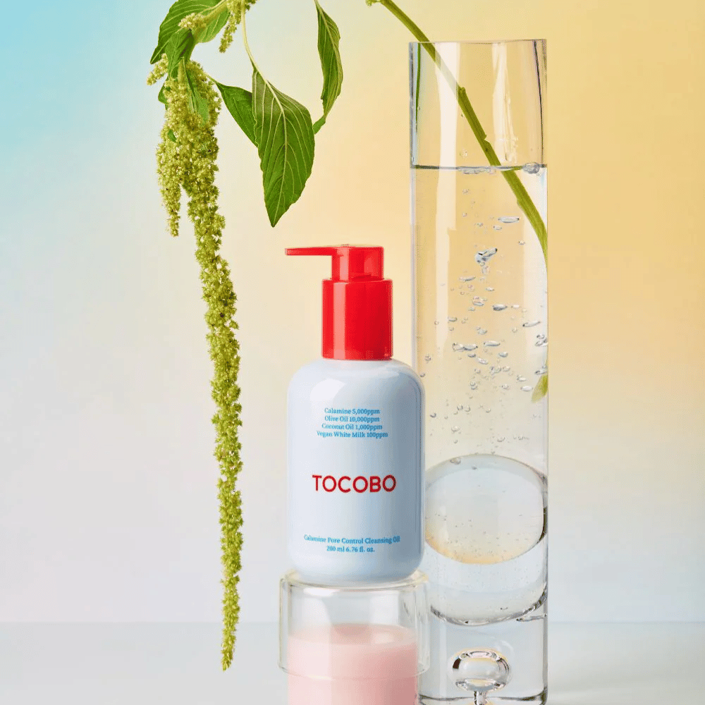 TOCOBO Calamine Pore Control Cleansing Oil med rød pumpe står ved siden af et højt glasrør fyldt med vand og en grøn plante, mod en lys pastelfarvet baggrund.