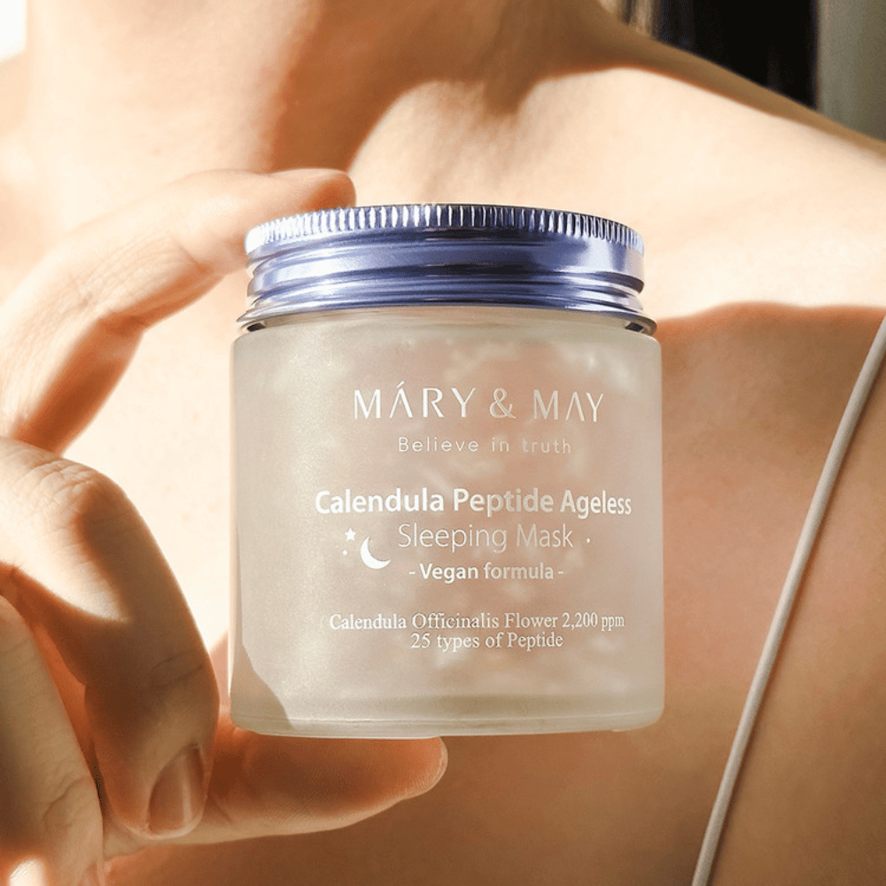 En kvinde holder en krukke med MARY & MAY Calendula Peptide Ageless Sleeping Mask. Krukken er gennemsigtig med et lilla låg, og teksten "Calendula Peptide Ageless Sleeping Mask" ses tydeligt. Produktet er en vegansk sovemaske med calendula og peptider, beregnet til at give huden en ungdommelig glød om natten.