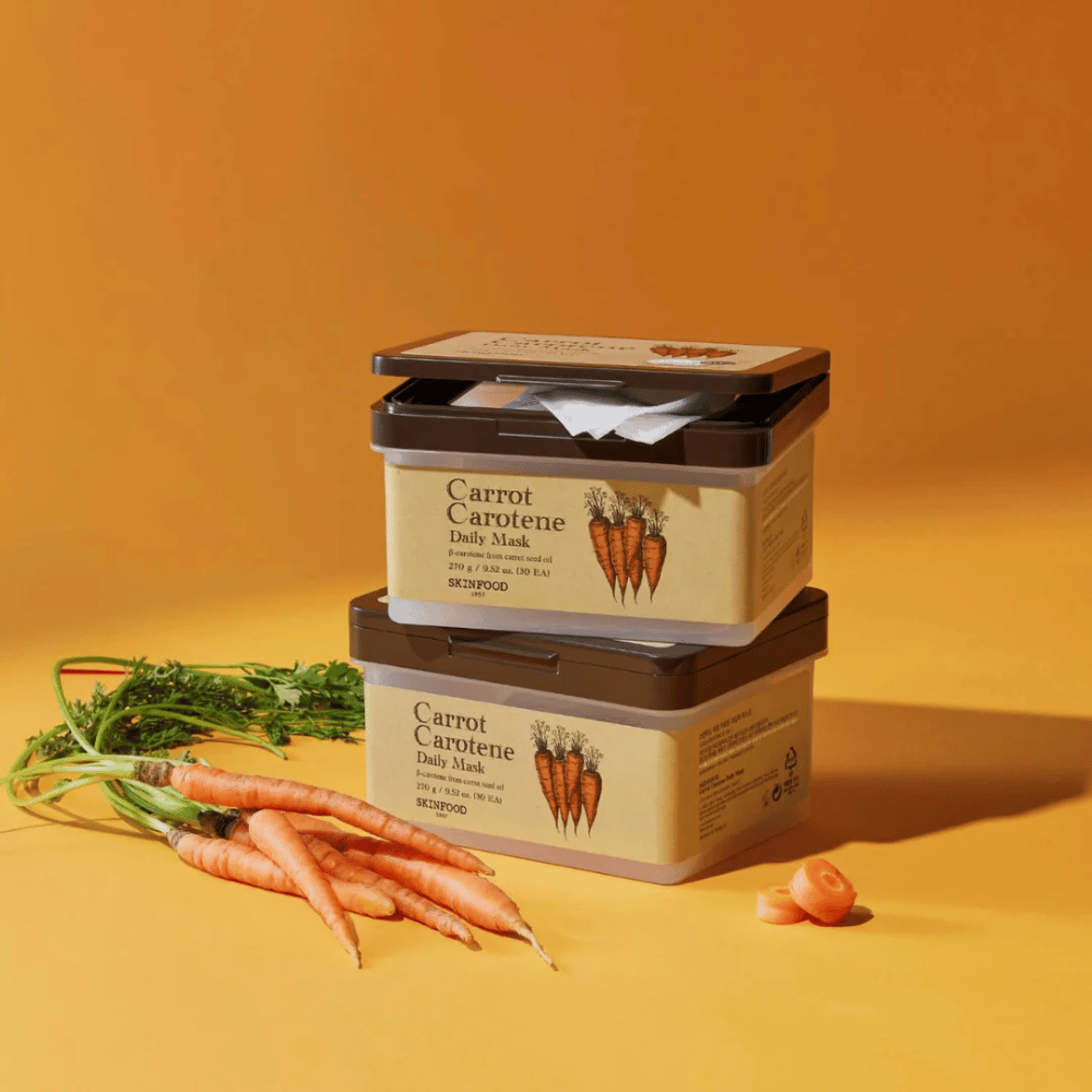 Billedet viser tre stablede pakker af "Carrot Carotene Daily Mask" fra SKINFOOD, placeret på en orange baggrund sammen med friske gulerødder. Hver kasse indeholder ansigtsmasker beriget med β-caroten fra gulerodsfrøolie, designet til daglig brug for at nære og pleje huden.
