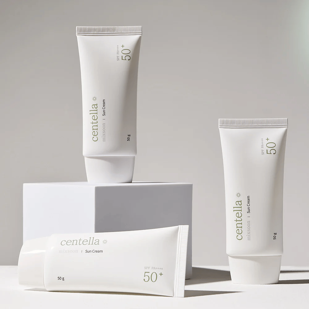 Tre pakker af Centella Sun Cream SPF 50+ PA++++ arrangeret æstetisk på hvide podier. To tuber står op og en ligger ned, alle med hvidt, minimalistisk design og grønne tekstdetaljer.